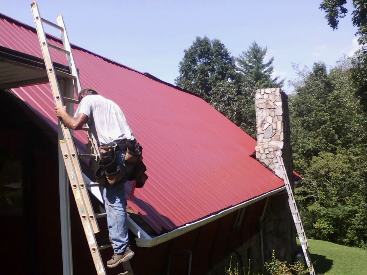 Roofer on a ladder beside a red metal roof and stone chimney — Soffit & Fascia Repair by Roofing Brothers in Lyman